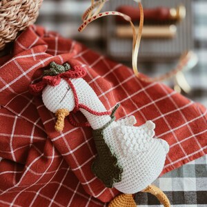 May include: A white crocheted goose toy featuring a yellow beak and feet, wearing a dark green leaf-shaped collar and a red flower-shaped hat with a thin red cord tied around its neck, resting on a red and white checkered fabric cloth.