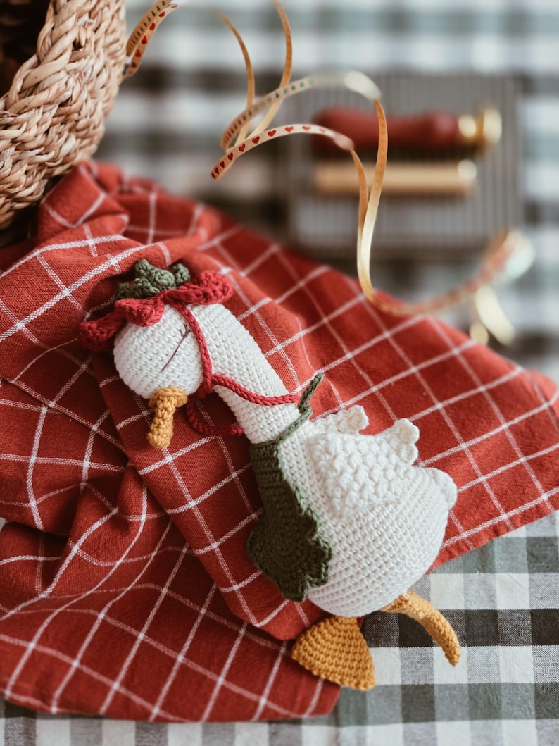May include: A white crocheted goose toy featuring a yellow beak and feet, wearing a dark green leaf-shaped collar and a red flower-shaped hat with a thin red cord tied around its neck, resting on a red and white checkered fabric cloth.