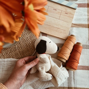 Op de afbeelding: Een gehaakte Snoopy-pop met zwarte en bruine details, een rode kraag en een wit lichaam. De afbeelding bevat een houten filmklapper, oranje garenklosjes en een beige en bruin geruite deken.