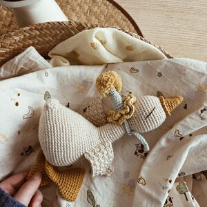 May include: A handmade crochet goose toy in cream, yellow, and blue. The goose has a yellow beak and feet, a ruffled collar, and a pom-pom on its head. It rests on a patterned blanket with illustrations of animals and suns.