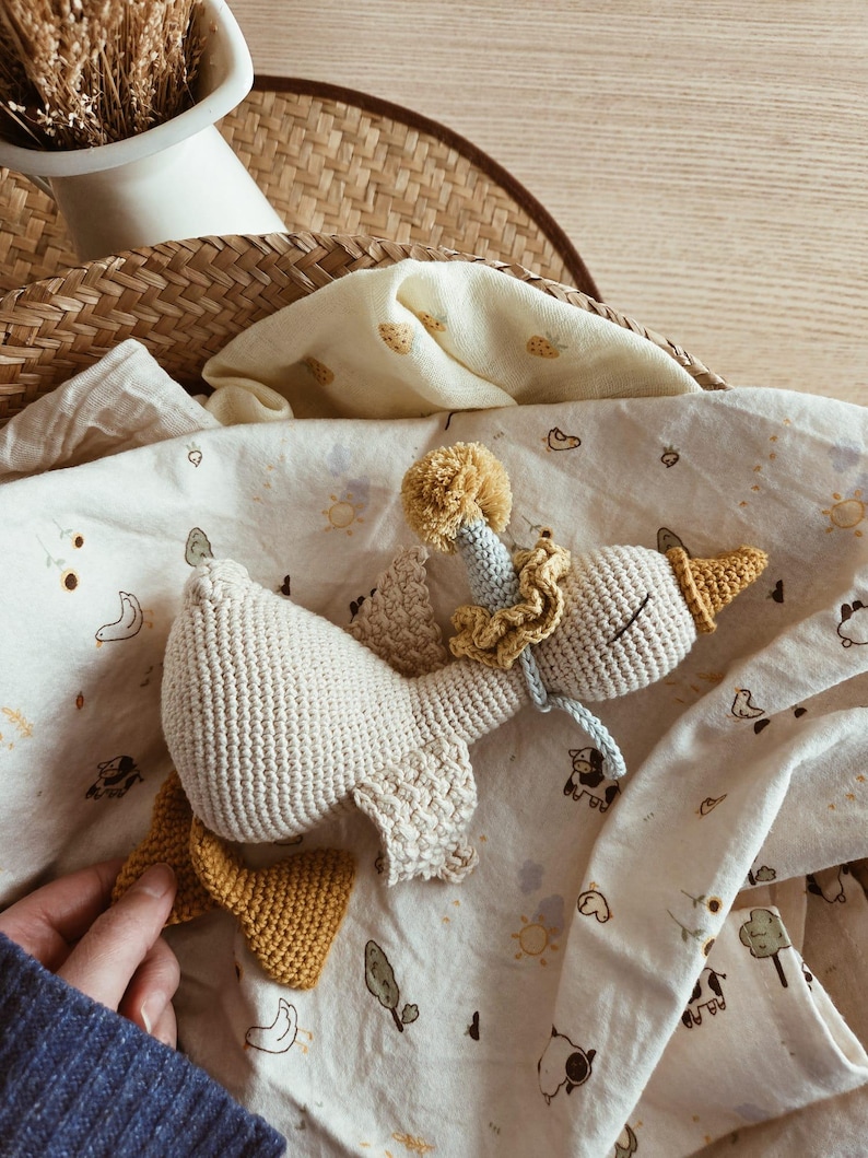 May include: A handmade crochet goose toy in cream, yellow, and blue. The goose has a yellow beak and feet, a ruffled collar, and a pom-pom on its head. It rests on a patterned blanket with illustrations of animals and suns.