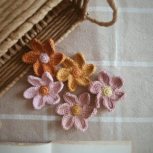 Pode incluir: Apresentação de flores de crochê feitas à mão em tons de laranja, amarelo e rosa. As flores estão dispostas sobre um tecido de tons neutros, com uma cesta de vime ao fundo. Um livro é parcialmente visível.