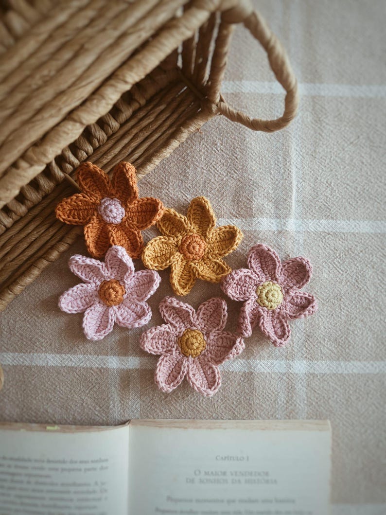May include: Close-up of several handmade crochet flowers in shades of orange, yellow, and pink. The flowers are arranged on a neutral fabric, with a woven basket in the background. A book is partially visible.