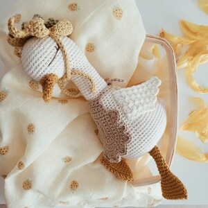 May include: A handmade crochet goose toy featuring a white body, a tan vest, and a yellow beak and feet. The goose has a small tan bonnet with a ruffled edge and is resting on a cream-colored muslin fabric patterned with small, light brown strawberry icons.