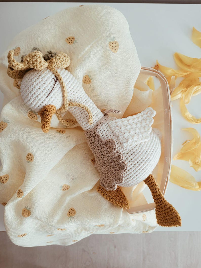 May include: A handmade crochet goose toy featuring a white body, a tan vest, and a yellow beak and feet. The goose has a small tan bonnet with a ruffled edge and is resting on a cream-colored muslin fabric patterned with small, light brown strawberry icons.