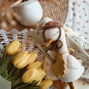 May include: A crocheted goose toy with a cream-colored body, wearing a brown hat with a small orange star, a mustard yellow scalloped bib, and featuring orange feet and a beak. The goose has closed eyes stitched in brown thread.