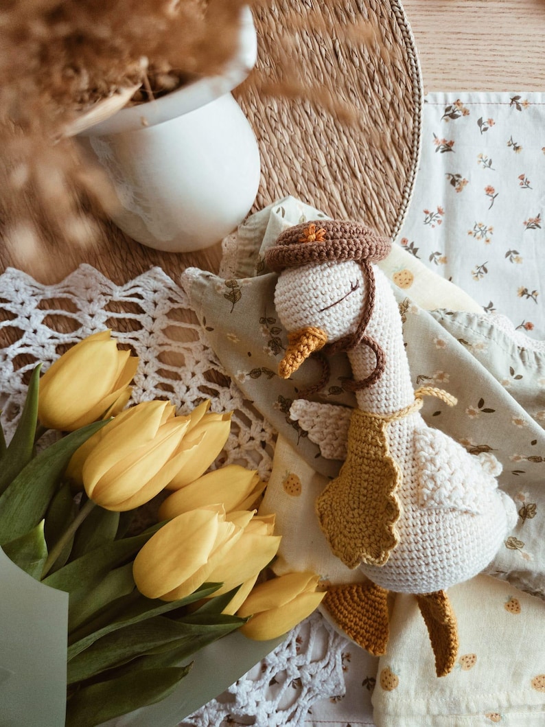 May include: A crocheted goose toy with a cream-colored body, wearing a brown hat with a small orange star, a mustard yellow scalloped bib, and featuring orange feet and a beak. The goose has closed eyes stitched in brown thread.