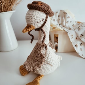Pode incluir: Uma boneca de ganso de crochê feita à mão em creme, marrom e amarelo. O ganso usa um chapéu e avental marrons, com pés e bico amarelos. A boneca está sentada, com os olhos fechados, e tem uma aparência caprichosa e artesanal.