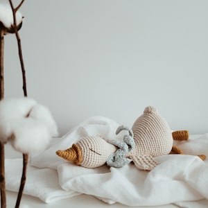 May include: Crocheted toys in the shape of a goose and a small bunny rest on a white surface. The goose is beige with a yellow beak and feet. A small blue bunny is nestled between the goose's head and body. The toys are on a white cloth.