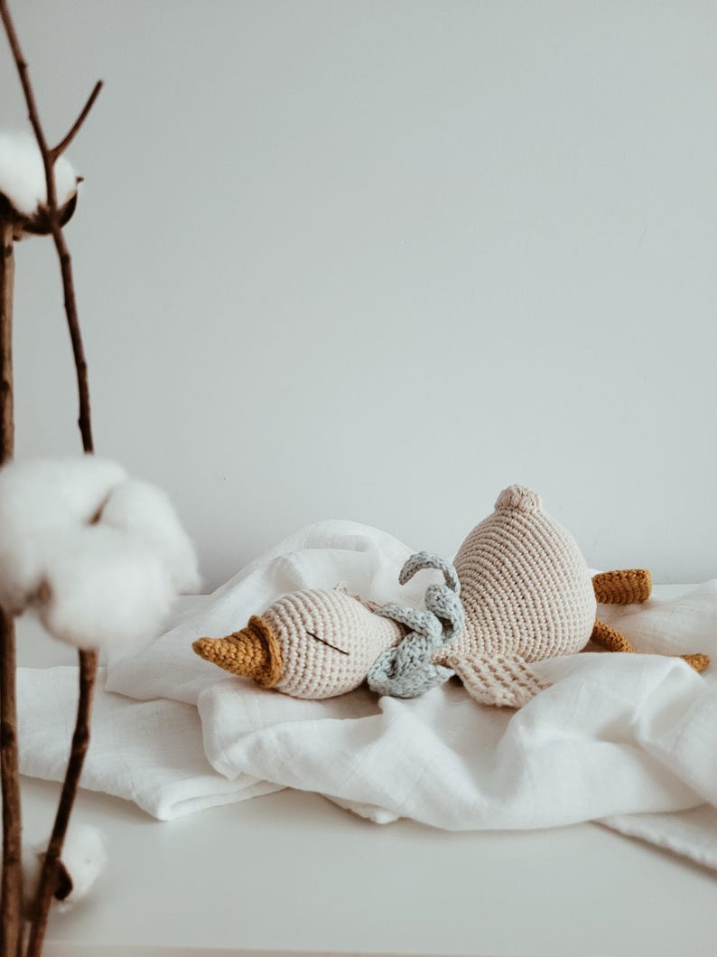 May include: Crocheted toys in the shape of a goose and a small bunny rest on a white surface. The goose is beige with a yellow beak and feet. A small blue bunny is nestled between the goose's head and body. The toys are on a white cloth.