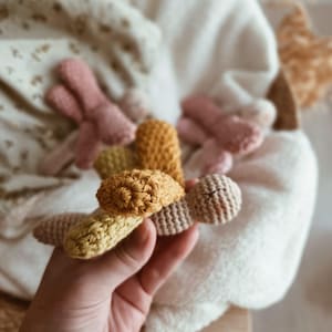 Pode incluir: Uma mão segura vários brinquedos de crochê em tons de amarelo, bege e rosa. Os brinquedos têm a forma de pequenos animais, com detalhes texturizados. Outros brinquedos semelhantes estão em uma cesta ao fundo, com um cobertor branco e macio.