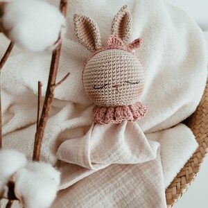 May include: A crocheted bunny baby lovey in beige and pink. The bunny has closed eyes, long ears, and a pink bow. The lovey is on a soft, cream-colored blanket, with a light pink muslin cloth attached.