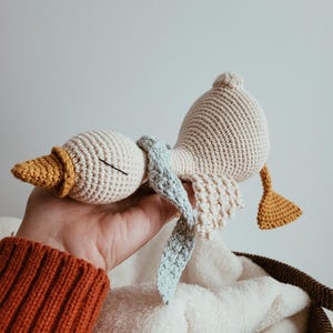May include: A handmade crochet goose toy in cream, yellow, and light blue. The goose has a textured body, a yellow beak, and a light blue scarf. The wings and tail are also yellow. The toy is resting on a white blanket.