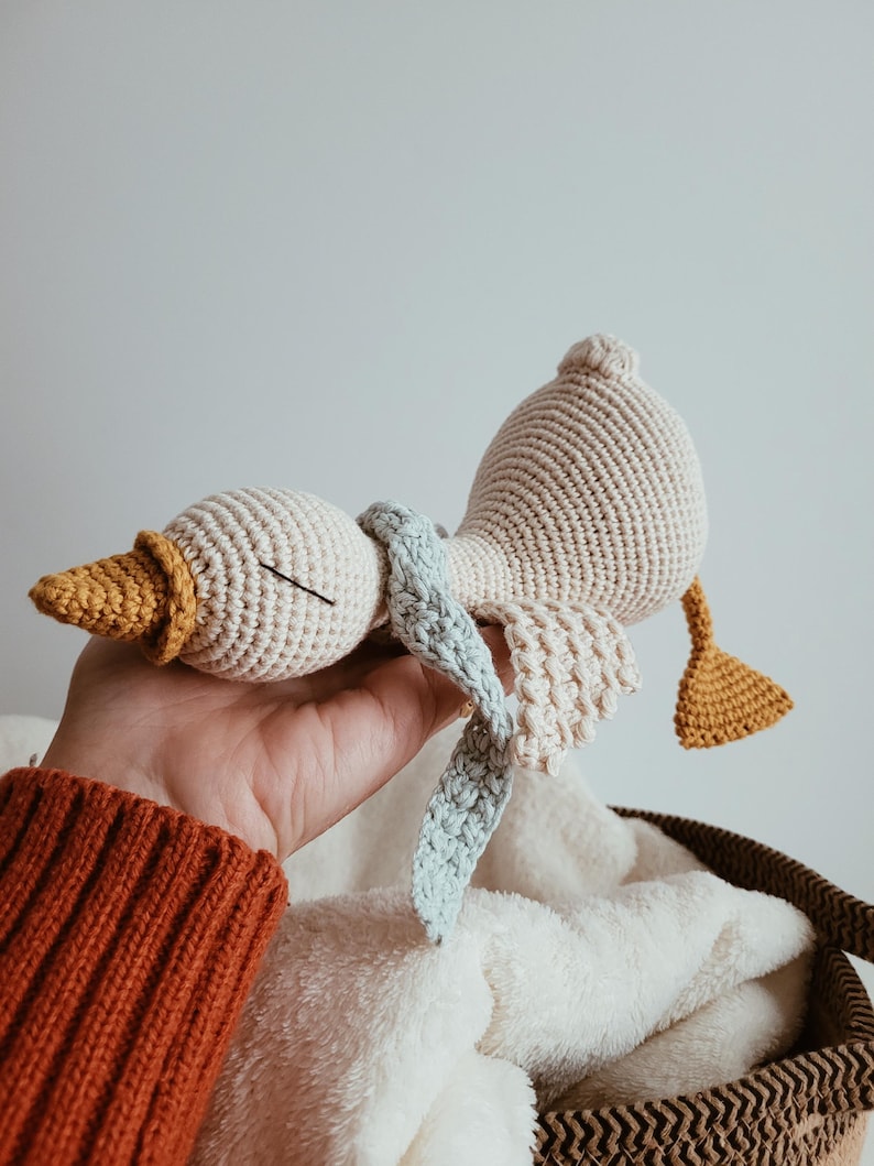 May include: A handmade crochet goose toy in cream, yellow, and light blue. The goose has a textured body, a yellow beak, and a light blue scarf. The wings and tail are also yellow. The toy is resting on a white blanket.
