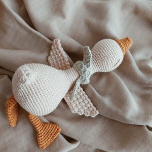 May include: A handmade crochet goose toy in cream, with orange feet, beak, and wing accents. The goose has a light green scarf and is lying on a beige fabric. This soft toy is perfect for a baby or child.
