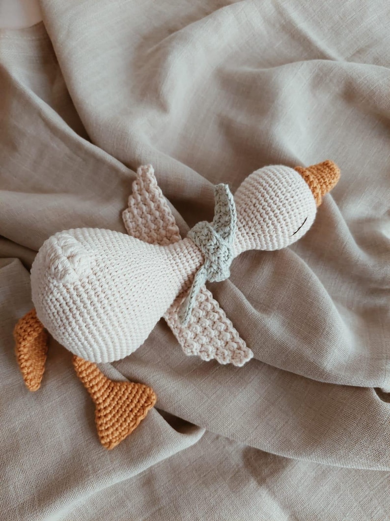 May include: A handmade crochet goose toy in cream, with orange feet, beak, and wing accents. The goose has a light green scarf and is lying on a beige fabric. This soft toy is perfect for a baby or child.