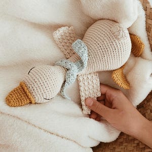 May include: A handmade crochet duck toy with a cream-colored body, yellow beak and feet, and a light blue bow. The duck is lying on a soft, white blanket inside a woven basket. A hand is holding the toy.