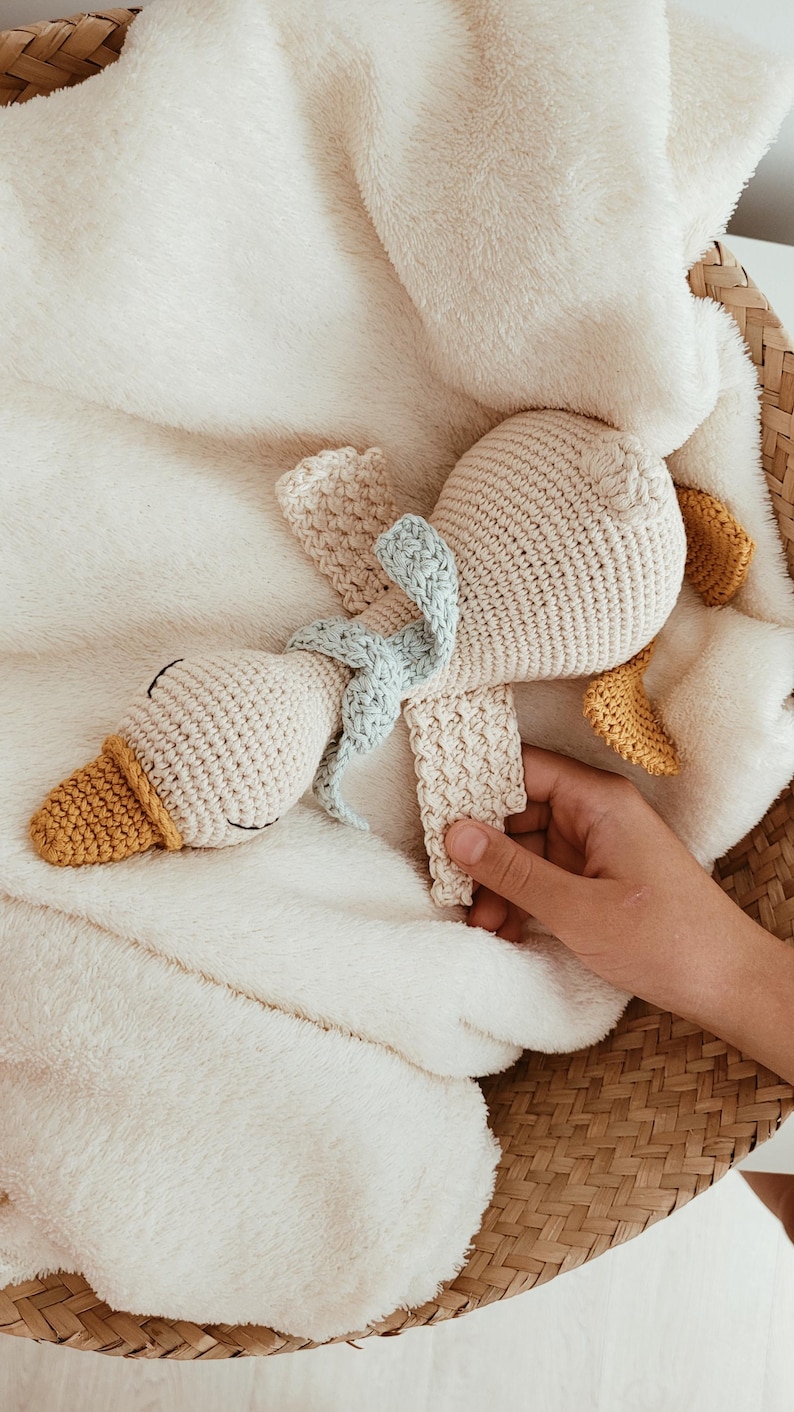 May include: A handmade crochet duck toy with a cream-colored body, yellow beak and feet, and a light blue bow. The duck is lying on a soft, white blanket inside a woven basket. A hand is holding the toy.