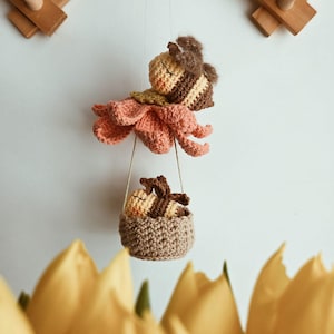 Pode incluir: Uma abelha de crochê feita à mão com um corpo em forma de flor e uma pequena cesta. A abelha é marrom e amarela, com um corpo de flor cor de pêssego. A cesta é marrom e contém outra abelha. O fundo é branco com detalhes em madeira.