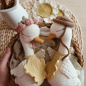 May include: Two handmade crochet geese with white bodies and orange beaks. One goose wears a pink flower crown and a yellow bib, while the other wears a brown hat and a yellow bib. Both have closed eyes and are positioned facing each other.