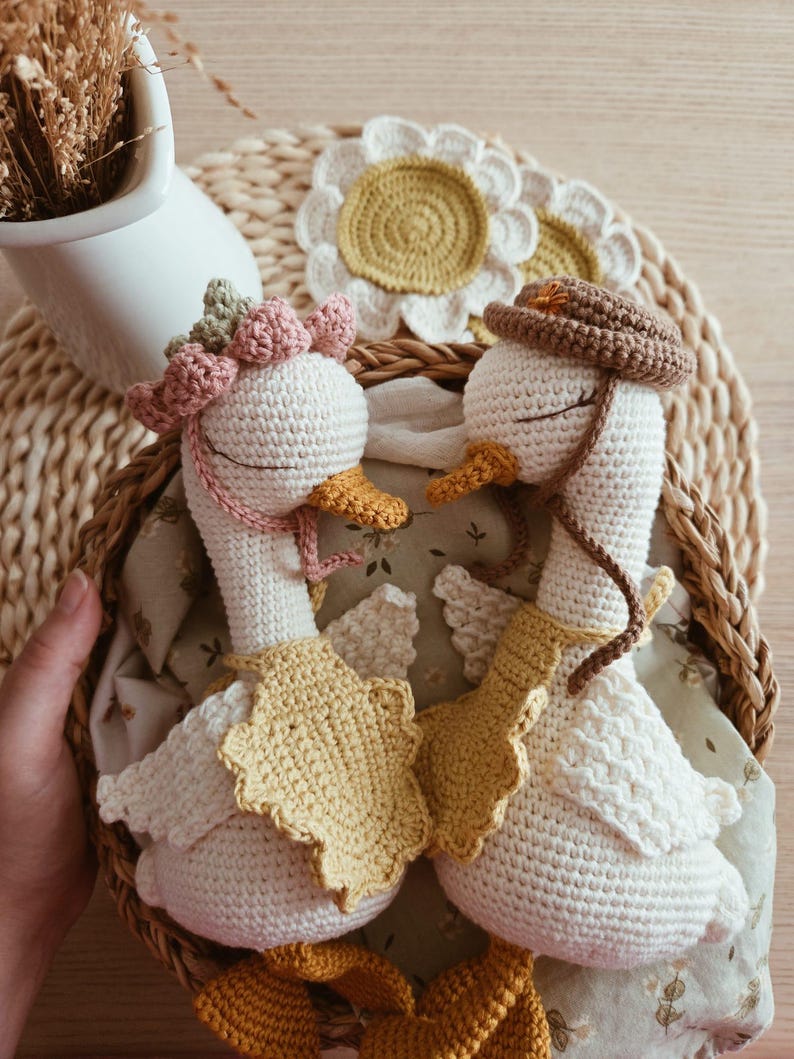May include: Two handmade crochet geese with white bodies and orange beaks. One goose wears a pink flower crown and a yellow bib, while the other wears a brown hat and a yellow bib. Both have closed eyes and are positioned facing each other.