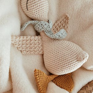 May include: A handmade crochet goose toy with a light beige body, a mustard yellow hat and feet, and a light blue scarf. The goose is lying on a soft, cream-colored blanket, creating a cozy and inviting scene.