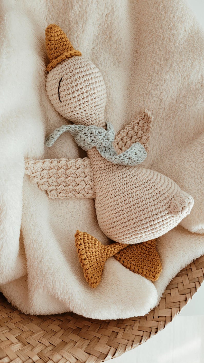 May include: A handmade crochet goose toy with a light beige body, a mustard yellow hat and feet, and a light blue scarf. The goose is lying on a soft, cream-colored blanket, creating a cozy and inviting scene.