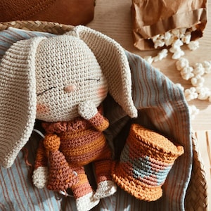 May include: A handmade crochet bunny doll with long ears, wearing an orange and brown outfit, and a matching hat. The doll is in a woven basket with a blue and white striped cloth. A brown leather bag and a paper bag of popcorn are in the background.