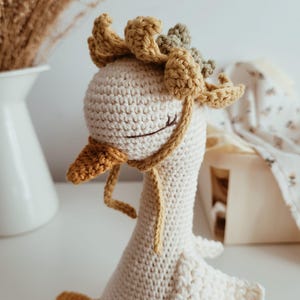 May include: A cream-colored crocheted goose toy featuring a mustard yellow beak and feet. The goose wears a mustard yellow crocheted bonnet with a ruffled edge and thin ties. The eyes are stitched in a closed, sleeping position with dark thread.