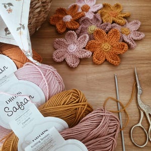 May include: A selection of crochet flowers in orange, pink, and yellow, with balls of yarn, a crochet hook, and vintage scissors. The yarn labels read "DROPS Safran". The items are arranged on a wooden surface, indicating a crafting project in progress.