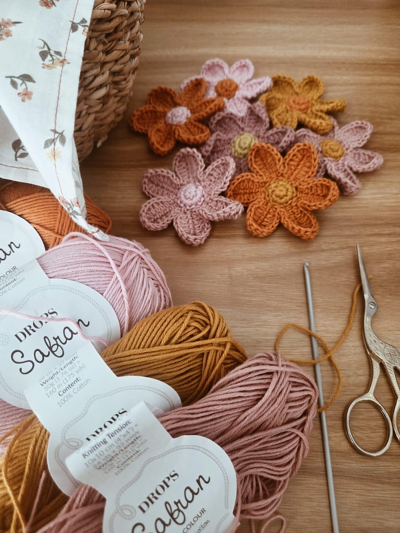 May include: A selection of crochet flowers in orange, pink, and yellow, with balls of yarn, a crochet hook, and vintage scissors. The yarn labels read "DROPS Safran". The items are arranged on a wooden surface, indicating a crafting project in progress.