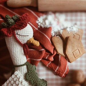 May include: A cream-colored crocheted goose toy featuring a dark green and red headpiece, a yellow beak, and a dark green scarf. The goose is positioned near a wooden rolling pin, a red and white striped cloth, and several light brown gingerbread cookies.