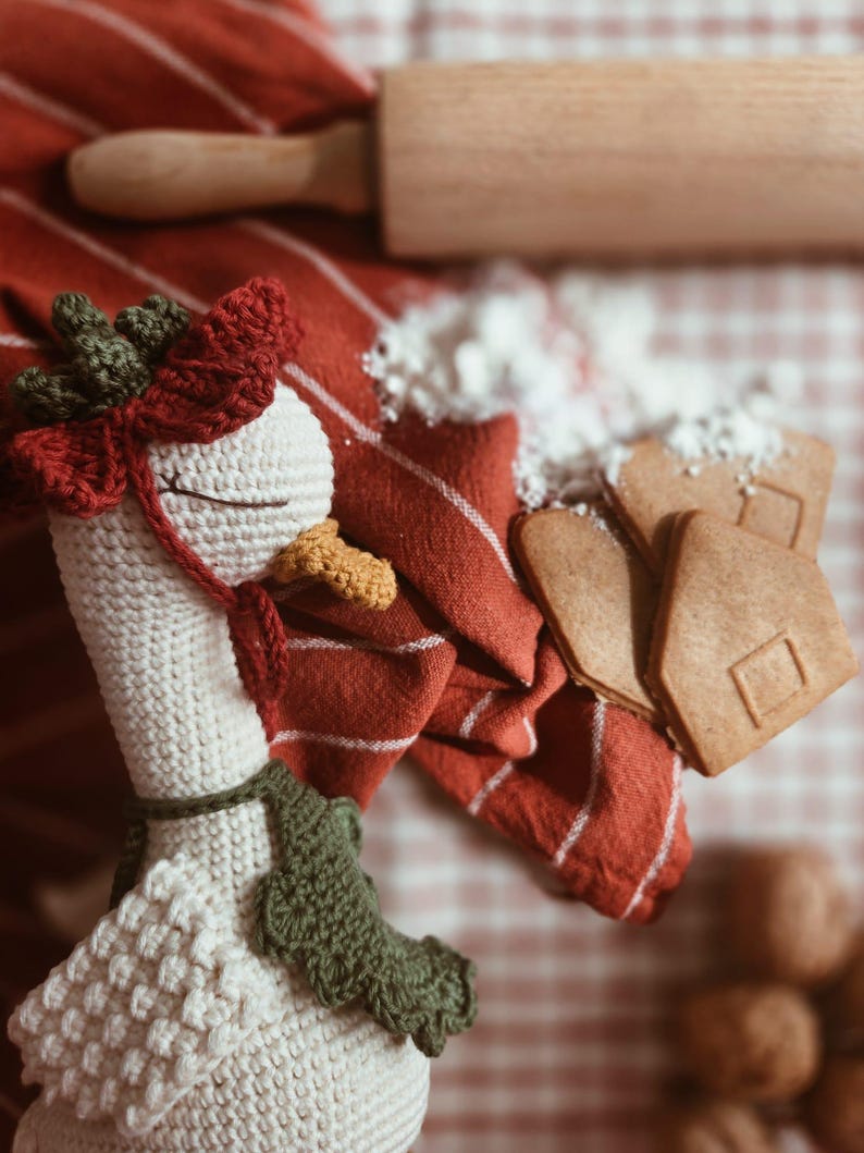 May include: A cream-colored crocheted goose toy featuring a dark green and red headpiece, a yellow beak, and a dark green scarf. The goose is positioned near a wooden rolling pin, a red and white striped cloth, and several light brown gingerbread cookies.