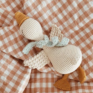 May include: A crocheted goose toy in cream, light blue, and mustard yellow. The handmade goose has a hat, scarf, wings, and feet. It is lying on a brown and white checkered blanket. The toy is soft and appears to be made of cotton.