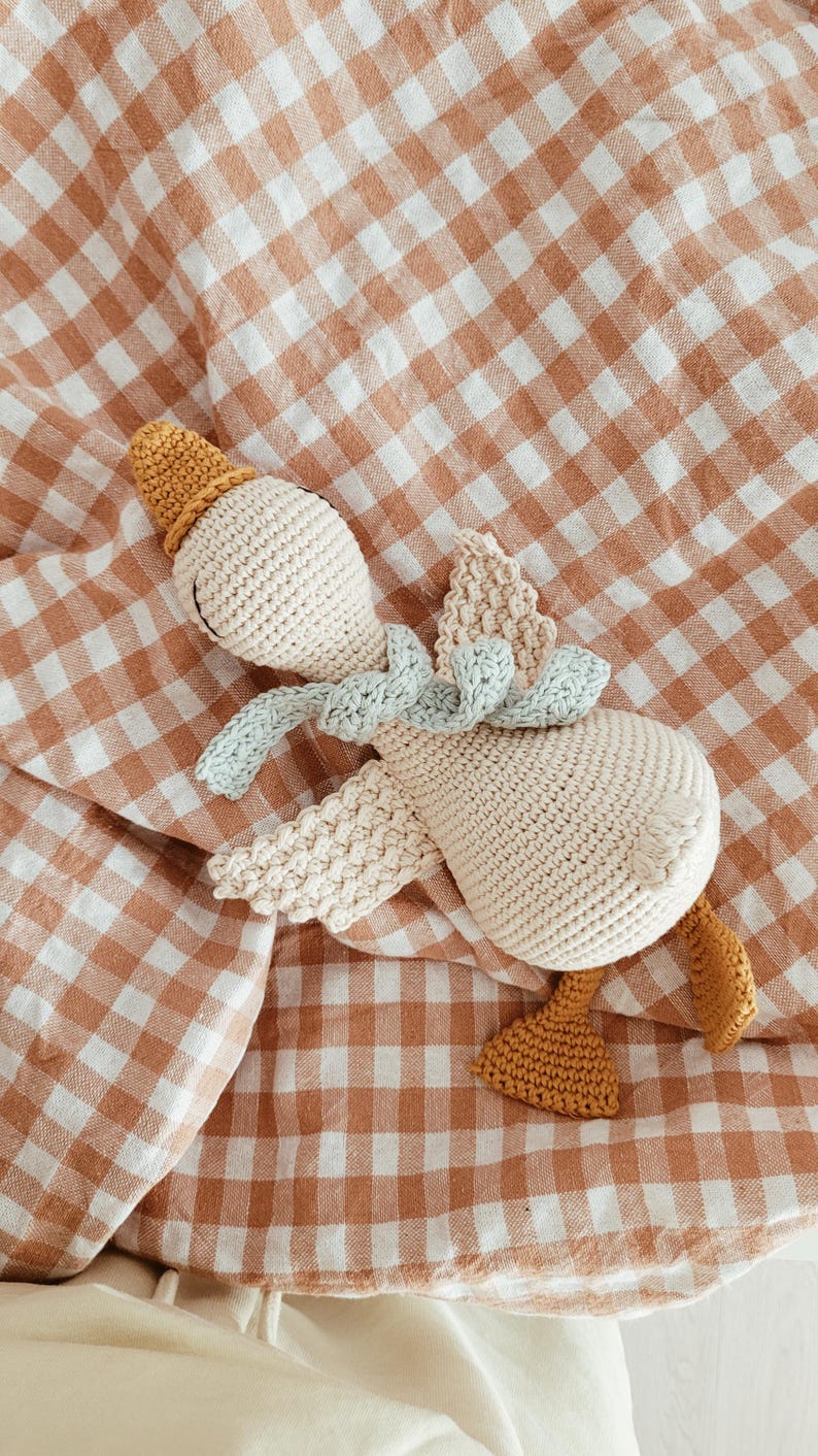 May include: A crocheted goose toy in cream, light blue, and mustard yellow. The handmade goose has a hat, scarf, wings, and feet. It is lying on a brown and white checkered blanket. The toy is soft and appears to be made of cotton.