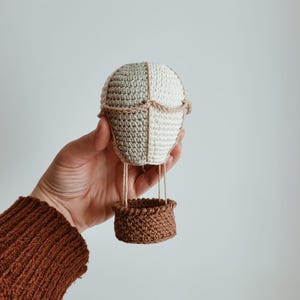 Puede incluir: Un globo aerostático de ganchillo hecho a mano en tonos beige, verde y marrón. El globo está dividido en cuatro secciones, con una cesta marrón suspendida debajo. El globo mide aproximadamente 13 cm de alto.