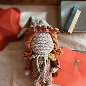 Pode incluir: Uma boneca de crochê com pele clara, cabelo laranja e um chapéu pontudo verde-sálvia. A boneca veste uma roupa estampada em verde-sálvia e laranja com gola de babados e segura um pequeno carimbo de lacre de madeira com cabeça metálica.
