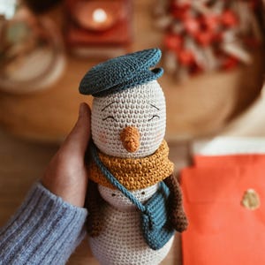 Op de afbeelding: Een gehaakt sneeuwpopfiguurtje met een lichtbeige lichaam, een donkerblauwe platte pet, een mosterdgele sjaal en een bijpassende donkerblauwe schoudertas. De sneeuwpop heeft een oranje wortelneus en geborduurde, gesloten ogen met een glimlach.