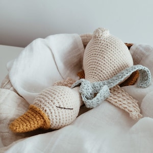 Pode incluir: Um brinquedo de ganso de crochê feito à mão em uma cesta. O ganso tem um corpo creme, um bico amarelo e um cachecol azul claro. O brinquedo está sobre um pano branco.