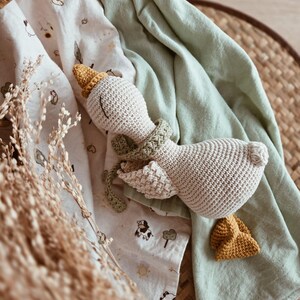 May include: A handmade, beige crochet goose toy with a yellow beak and hat, and a green scarf. The toy rests on a light green blanket and a patterned white blanket with animal illustrations. The background includes a woven basket and dried plants.