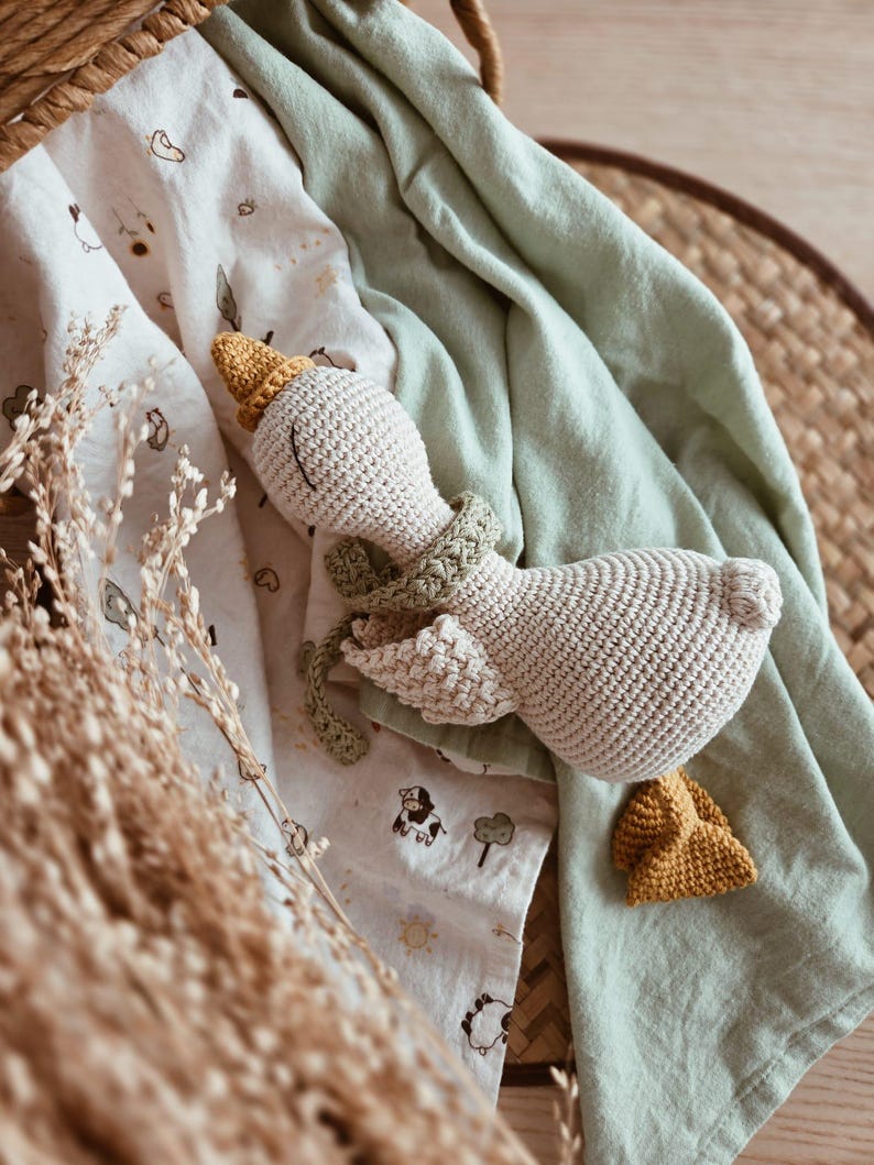 May include: A handmade, beige crochet goose toy with a yellow beak and hat, and a green scarf. The toy rests on a light green blanket and a patterned white blanket with animal illustrations. The background includes a woven basket and dried plants.