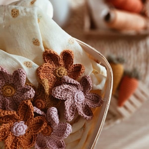 May include: A wooden basket contains crocheted flowers in shades of orange and pink, set on a pale yellow fabric with strawberry motifs. The flowers are the main focus, displaying intricate crochet detail.