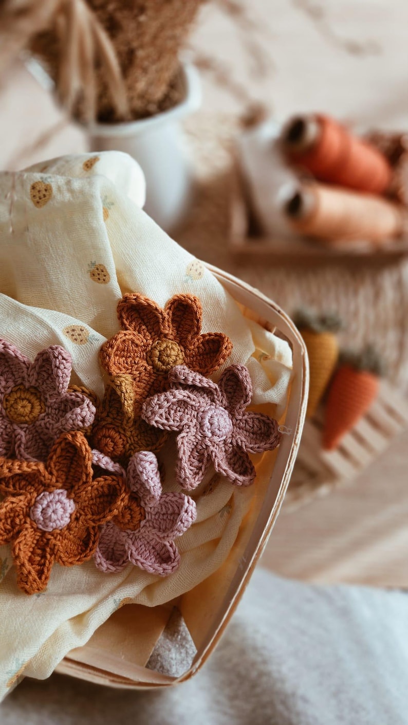 May include: A wooden basket contains crocheted flowers in shades of orange and pink, set on a pale yellow fabric with strawberry motifs. The flowers are the main focus, displaying intricate crochet detail.