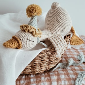 May include: A handmade crochet goose doll with a party hat and a decorative collar, resting in a woven basket. The goose is cream-colored with a yellow beak and accents. The basket sits on a checkered fabric.