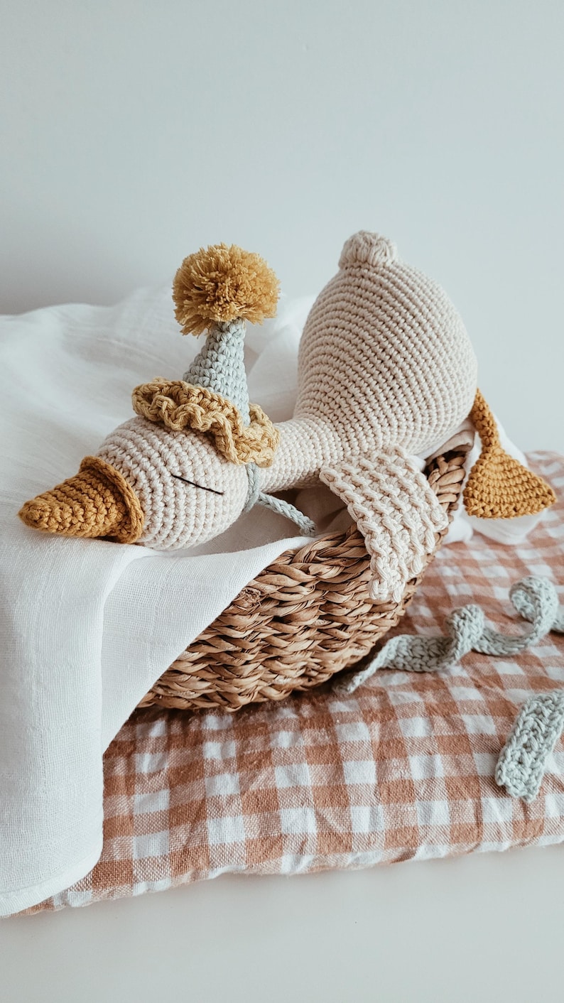 May include: A handmade crochet goose doll with a party hat and a decorative collar, resting in a woven basket. The goose is cream-colored with a yellow beak and accents. The basket sits on a checkered fabric.