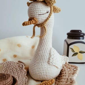 May include: A handcrafted crochet goose toy in cream yarn with a long neck, closed eyes, and a textured orange beak. The goose wears a decorative mustard yellow bonnet with ruffled edges and ties. Beside it are small crocheted accessories in brown.