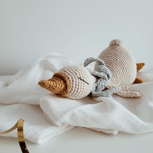 May include: A crocheted goose toy in cream, tan, and mustard yellow. The handmade plush animal has a long neck and a beak. The goose is lying on a white cloth. A gold ribbon is in the foreground.