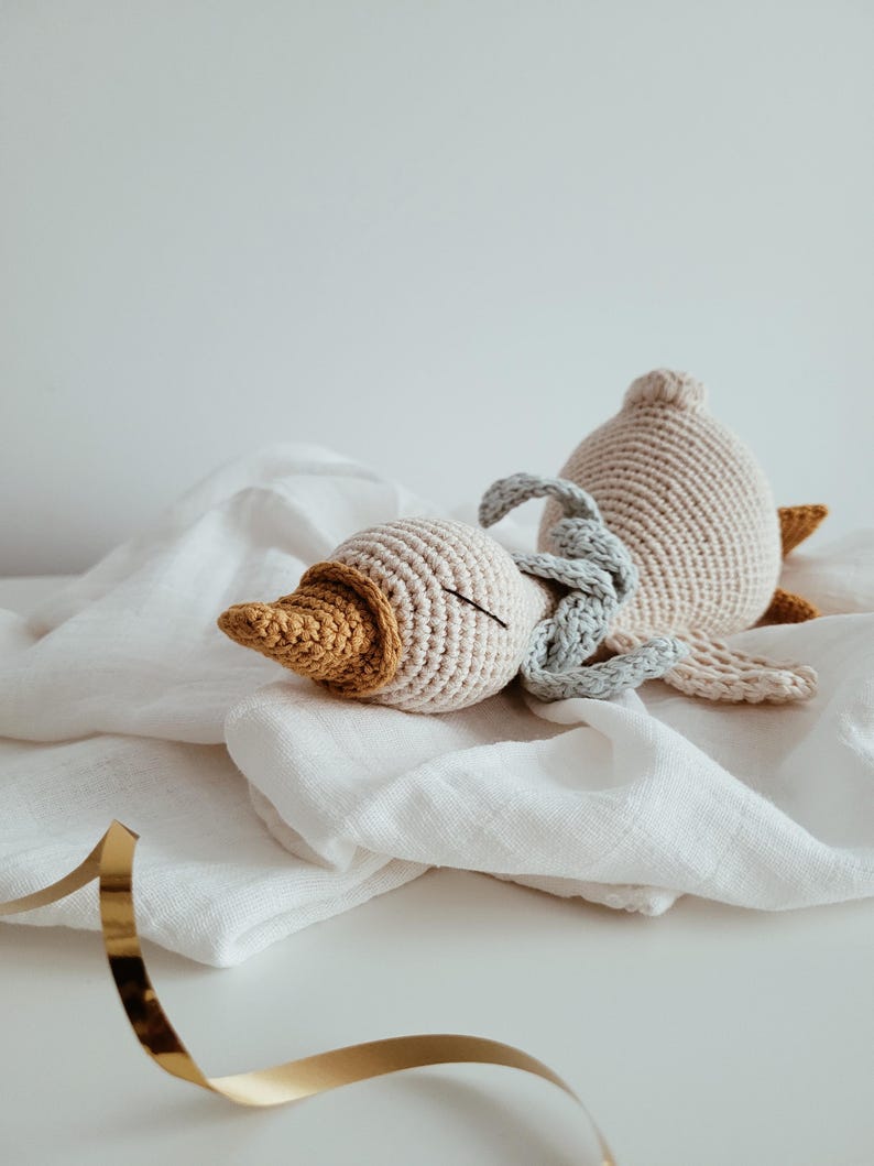 May include: A crocheted goose toy in cream, tan, and mustard yellow. The handmade plush animal has a long neck and a beak. The goose is lying on a white cloth. A gold ribbon is in the foreground.