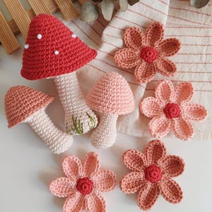 May include: A selection of handmade crochet items, featuring three mushroom figures and several flower appliqu&eacute;s. The mushrooms have red, pink, and white caps, with white stems. The flowers are pink and coral, with red centres. The items are arranged on a white surface.