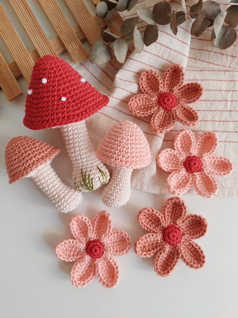 May include: A selection of handmade crochet items, featuring three mushroom figures and several flower appliqu&eacute;s. The mushrooms have red, pink, and white caps, with white stems. The flowers are pink and coral, with red centres. The items are arranged on a white surface.